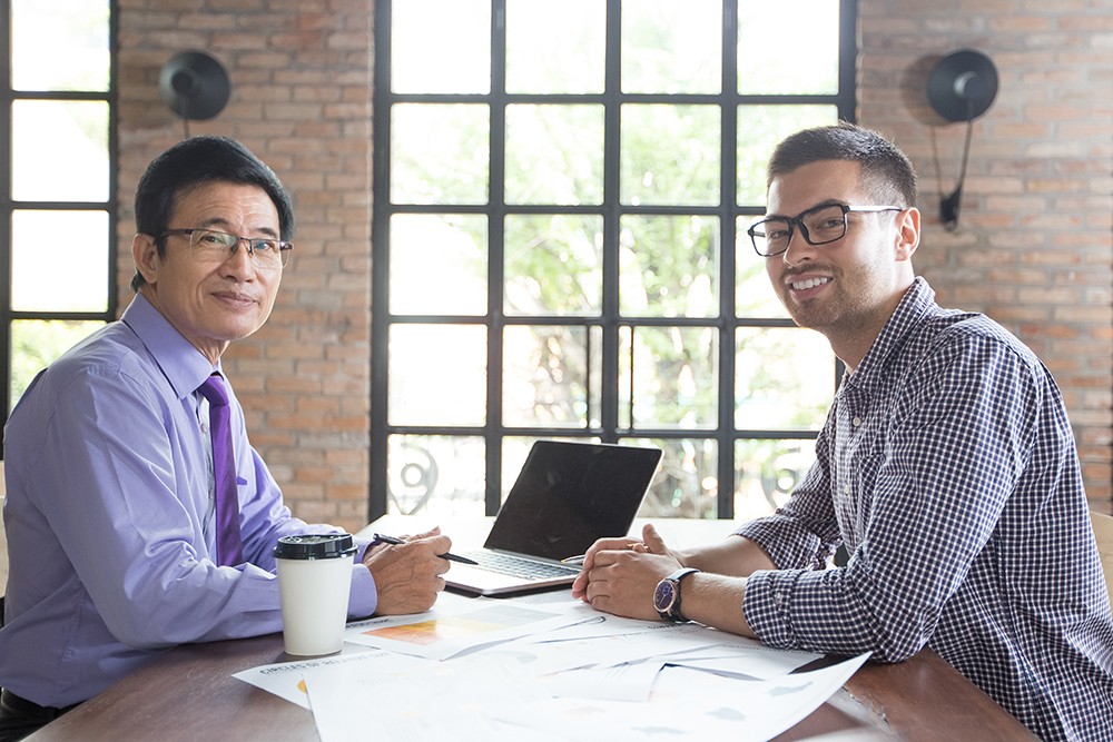 Closeup of two smiling business men looking at camera, discussing issues and working at office desk with brick walls and loft windows in background. Side view.